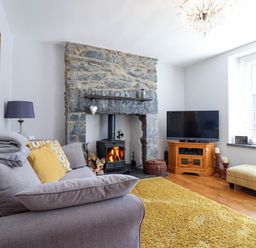 A living room with a stone fireplace wood stove grey sofa yellow rug and a television at Dola in Pentre Du near Betws-Y-Coed