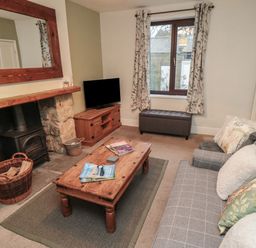 A living room with a wooden coffee table magazines on it a gray sofa cushions a fireplace with a wood-burning stove a wicker basket with logs a wooden TV cabinet a window with floral curtains and a gray ottoman at Providence Cottage in Ugglebarnby near Sleights