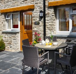 An outdoor patio with a round table set with cups and juice and four wicker chairs in front of a stone wall house with wooden door and two windows at Teifi Cottage in Cardigan Near Cenarth