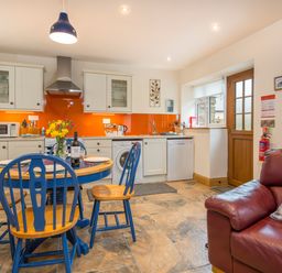 A kitchen with blue and wood dining chairs and table a red leather sofa and orange backsplash at Cych Cottage in Cardigan Near Cenarth