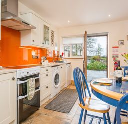A kitchen with white cabinets orange backsplash and blue dining table with chairs and flowers at Cych Cottage Cardigan Near Cenarth