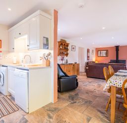 A kitchen with white cabinets and appliances next to a dining area with a wooden table and chairs and a living room with leather sofas and a wood stove at Cothi Cottage in Cardigan Near Cenarth