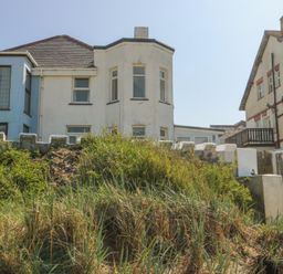 A two story white house with bay windows behind a white fence and overgrown grass at The Harbour in Rhosneigr