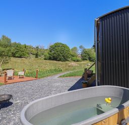 An outdoor area with a hot tub containing a rubber duck next to a black corrugated building and a deck with two chairs at Celyn in Bontddu near Dolgellau