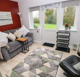 A sitting room with a gray sofa with cushions a small wooden side table a patterned rug a black leather chair a shoe rack and a door leading to a garden at Orchard View in Pembroke