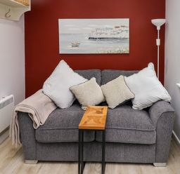 A gray sofa with four cushions and a beige throw blanket a small wooden table a floor lamp a painting on a red wall a window with a green vase and a small wooden house ornament at Orchard View in Pembroke