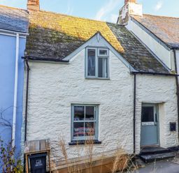 A white stone cottage with a mossy roof two windows a blue door and a wooden bench outside at Alpha Cottage in Dittisham near Dartmouth