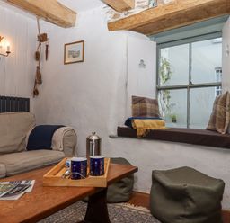 A sitting area with a sofa a wooden table with cups a window seat with cushions and poufs at Alpha Cottage in Dittisham near Dartmouth