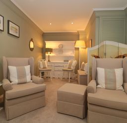 A sitting room with two beige armchairs with cushions a beige ottoman a white side table two white chairs a white cabinet with shelves and lamps at Tree Tops in Broadway