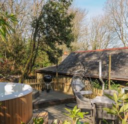 An outdoor wooden deck with a round hot tub a patio table with four chairs a hanging chair with cushions and a barbecue grill at Rose Cottage in Callington