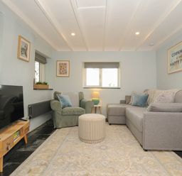 A living room with a grey sofa, green armchair, round ottoman, wooden TV stand, and framed art at Rose Cottage in Callington