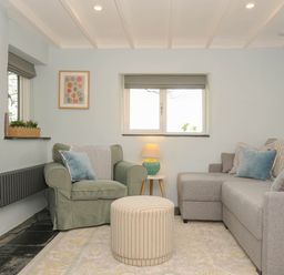 A living room with a green armchair a gray sofa a striped ottoman and a lamp on a small wooden table at Rose Cottage in Callington