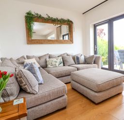 A living room with a gray sectional sofa and matching ottoman by glass doors with a wooden side table with red tulips and a large mirror above the sofa at Buzzards View in Dyserth