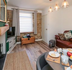 A living room with a dining table set with cups and eggs a brown leather sofa a television on a stand and a wooden chest by the window at Number Ten in Amble