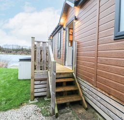 Wooden steps leading to an open door of a brown cabin near a lake with grass and gravel pathway at 12A Lake Escape in Carnforth