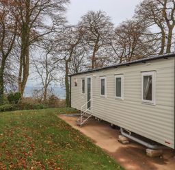 A mobile home on a grassy area with fallen leaves near trees and a body of water at 5 Manor Park in Bucks Cross Bideford
