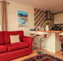 A living room with a red sofa and beige pillows next to a kitchen island with three white bar stools a tiled backsplash and a vase of flowers on the counter at Garden View in Hillhead near Brixham