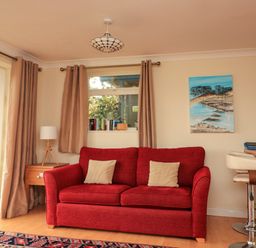 A living room with a red sofa two beige cushions a wooden side table with a lamp and a painting on the wall at Garden View Hillhead near Brixham