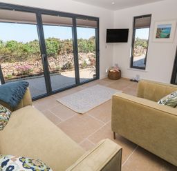 A sitting room with two beige sofas a small rug a wall mounted tv and large windows overlooking a garden at Little Victory in Pendeen