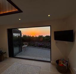 A room with a rug on the floor and a wall-mounted TV next to an open large door showing an outdoor patio at sunset at Little Victory in Pendeen
