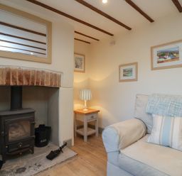A sitting room with a wood burning stove a small wooden side table lamp and an armchair with cushions at Curlew Cottage in North Curry near Taunton