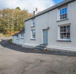 The exterior of a white house with multiple windows and a light blue door next to a paved driveway and a wooded area at Leeward Cottage in Cosheston near Pembroke Dock
