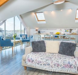 An open living area with a sofa and two blue chairs near large windows overlooking water at Leeward Cottage in Cosheston near Pembroke Dock