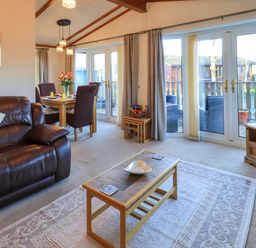 A living room with a brown leather sofa coffee table dining table with chairs and large windows at Lavender Lodge South Lakeland Leisure Village