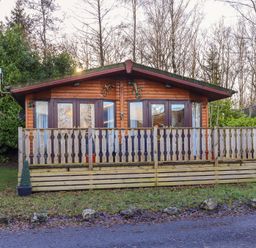A wooden cabin with a fenced porch and glass doors surrounded by trees at Lavender Lodge in South Lakeland Leisure Village