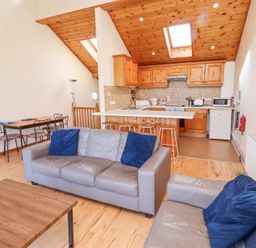 An open living room and kitchen area with gray sofas and blue pillows a wooden dining table with chairs and a kitchen with wooden cabinets and bar stools at Waters Edge in Embleton