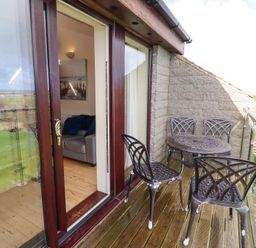 A balcony with four metal chairs and a round metal table overlooking a grassy area and a room with a sofa visible through sliding glass doors at Waters Edge in Embleton