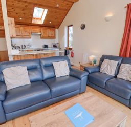 A living room with blue sofas and pillows near a wooden kitchen area with skylights at Dunstanburgh View in Embleton