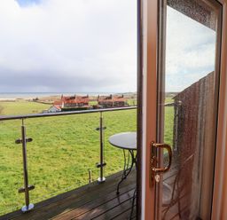 A balcony with a round table overlooking green fields and houses at Dunstanburgh View in Embleton