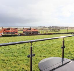 A balcony with glass railing overlooking green fields and houses with red roofs at Dunstanburgh View in Embleton