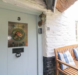 A wooden bench with striped cushions next to a white stone wall and a light blue door with stained glass at 3 Cumberland Cottages in Ringmore