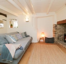 A living room with a gray sofa and cushions next to a stone fireplace with a fire burning at 3 Cumberland Cottages in Ringmore