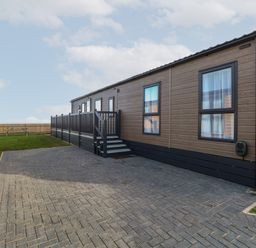 A driveway with a paved parking area next to a single story home with wooden siding and a fenced deck at Dorchester in Broomholm near Mundesley
