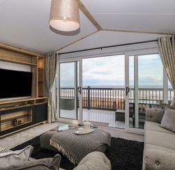 A living room with sofas a TV unit and sliding glass doors opening to a balcony with ocean view at Dorchester in Broomholm near Mundesley