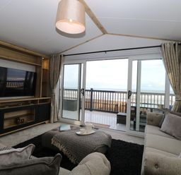A living room with sofas a TV cabinet and sliding glass doors opening to a balcony overlooking the sea at Dorchester in Broomholm near Mundesley