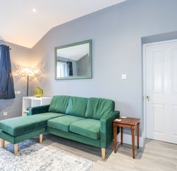A living room with a green sofa a white door a wooden side table a white shelf with a green vase a floor lamp and a mirror on a grey wall at The Fir Grove Annex in Menai Bridge