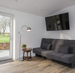 A sitting area with a gray couch, a floor lamp, a small wooden table with plants and cups, a wall-mounted TV, and a glass door showing an outdoor grassy area at Ynys Uchaf Near Benllech