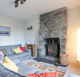 A living room with a stone fireplace a wood stove two grey sofas with colored cushions a small wooden bookshelf a tall floor lamp and a colorful rug at Gwelfor in Harlech