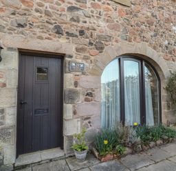 A stone exterior wall with a dark wooden door a lamp and an arched window with plants outside at Primula Patch Akeld near Wooler