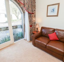A brown leather sofa with a red cushion near a window with floral curtains and a side table with a lamp at Primula Patch in Akeld near Wooler