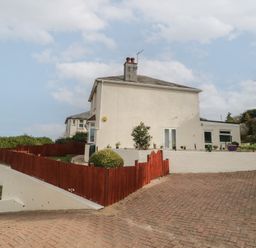 A paved driveway leading to a white two-story house with red wooden fencing and small garden plants at Hilltop Cottage in Paignton