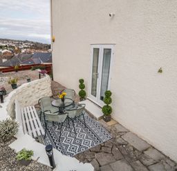 An outdoor patio area with a round glass table and four chairs on a patterned rug next to a white bench and potted plants at Hilltop Cottage in Paignton