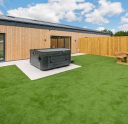 An outdoor area with a hot tub and wooden picnic table surrounded by artificial grass at Bears Court Barn 2 in Little Rissington near Bourton-On-The-Water