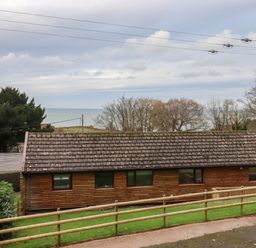 A wooden single-story building with a tiled roof and windows surrounded by grass and trees overlooking the sea at Castle View Lodge in Ilfracombe