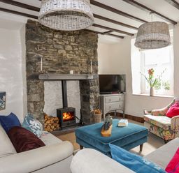 A living room with a stone fireplace and wood burner two sofas an ottoman and a floral armchair with a window at Riverwash Cottage in Dwygyfylchi