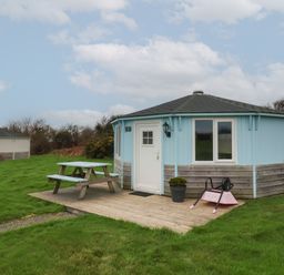 A small round blue and wood cabin with a picnic table and a pink wheelbarrow on a wooden deck surrounded by grass at Shipload Bay near Hartland